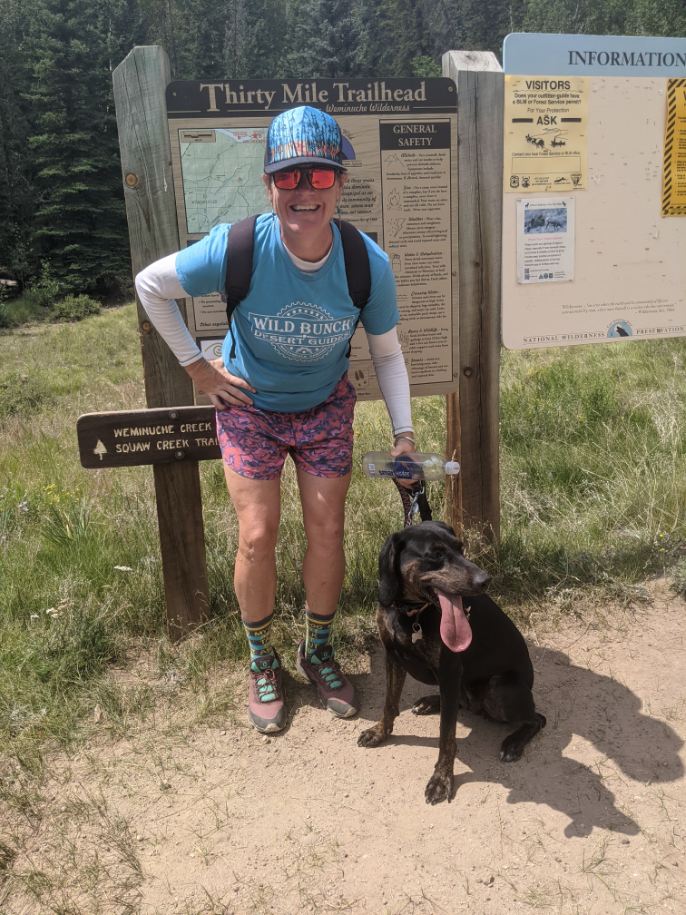 A beaming Laurel Darren takes a Lake City hiking break with her loyal sidekick Daisy Mae in front of the Thirty Mile Trailhead sign with the vast beauty of Colorado's San Juan Mountains behind them.