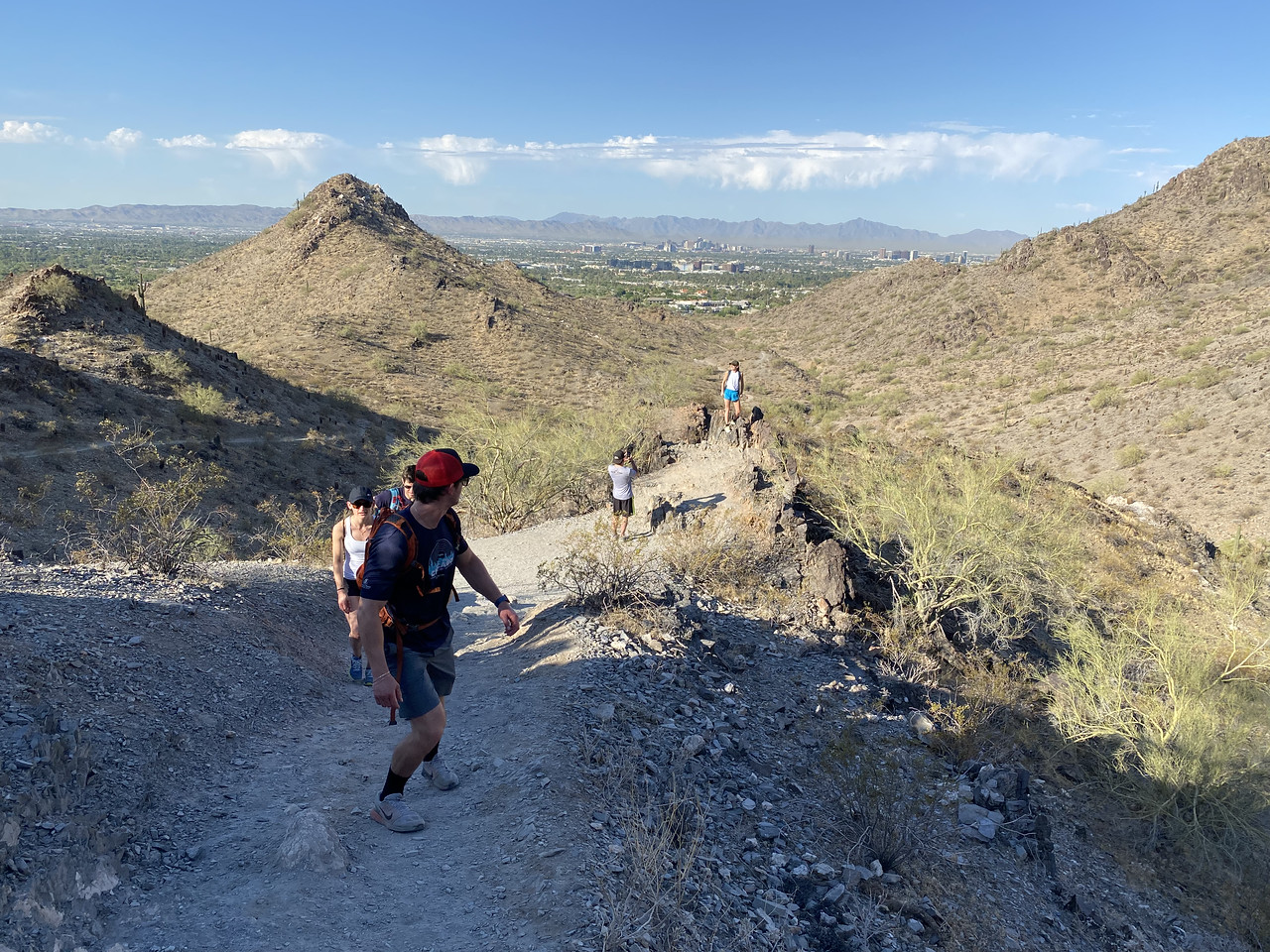 A hiking group makes its way up a scenic hill during a Wild Bunch Desert Guides outing. The leader of the pack (orange hat) peeks over his shoulder to admire the view and spies the last two hikers pausing to take a picture with the sweeping landscape behind them. 