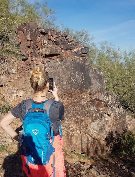 A female hiker pauses in front of a rock formation to take a photo of the Sonoran Desert scenery during a recent guided Phoenix hiking tour with the Wild Bunch Desert Guides.