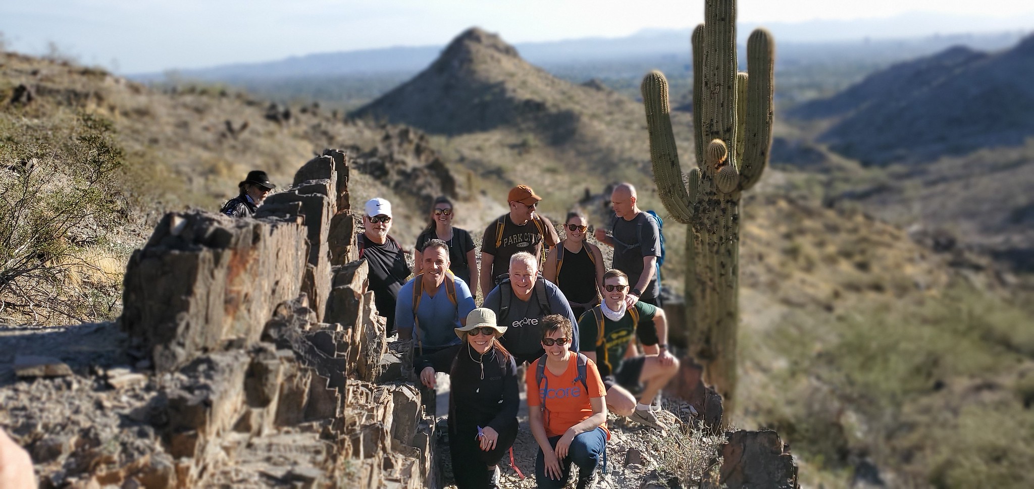 With an incredible desert vista as the backdrop, and a cactus looming over their shoulders, an early-morning summer hiking group pauses for a photo on a particularly picturesque part of their path.