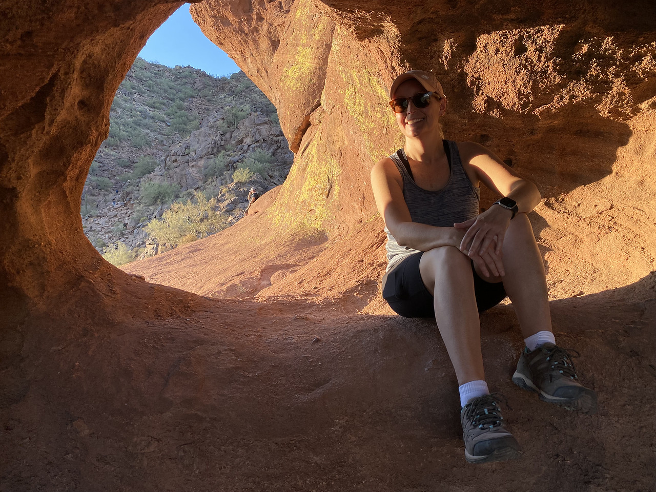 A woman is all smiles while sitting down to take a break after finding some shade in a natural tunnel in a rock formation during a Scottsdale hiking tour.