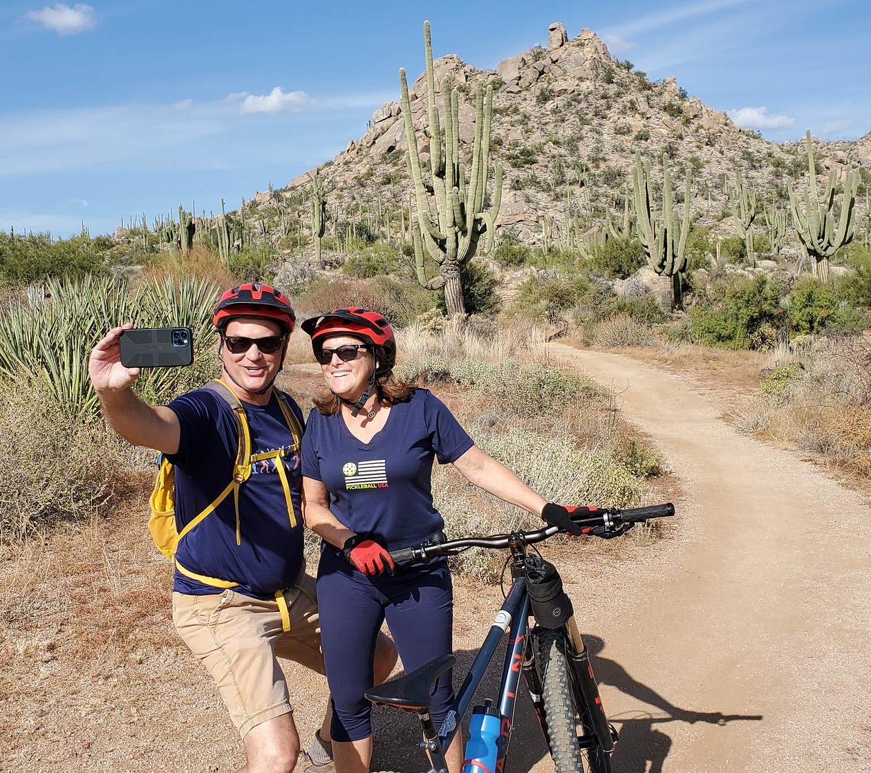 A married couple enjoying a Scottsdale mountain bike tour with the Wild Bunch Desert Guides pauses for a selfie together along the trail with a picturesque background behind them.