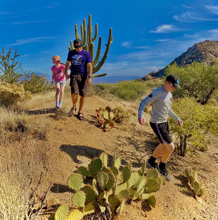 A father (center) enjoys some quality time with his children during a Wild Bunch Desert Guides hike in Phoenix AZ. While dad holds the hand of his daughter (left), they watch his son (right) descend a colorful Cactus dotted hill.