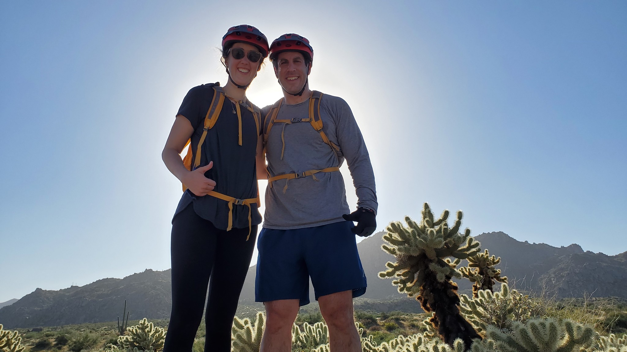 With the sun shining brightly behind them, and standing in the midst of the sticky Teddy Bear Cactus, a smiling couple are all thumbs up about their memorable Phoenix adventure tour with the Wild Bunch Desert Guides.