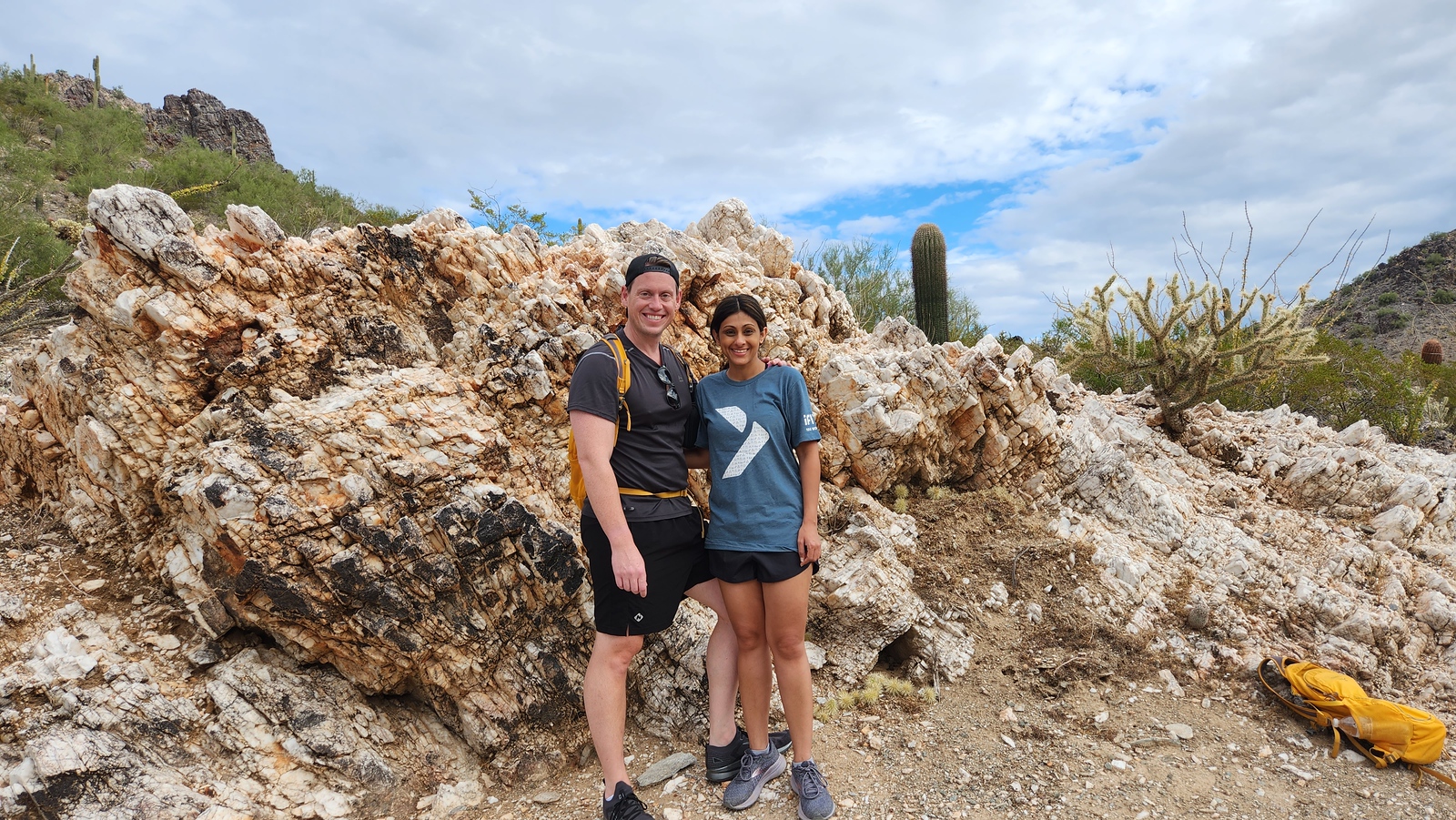 A couple is all smiles posing in front of a Sonoran Desert rock formation with a wide variety of cactus also in the picture.