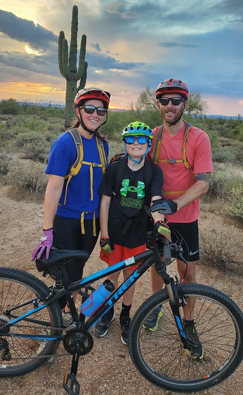 With a beautiful Arizona sunset as the backdrop, a smiling son and his beaming parents pose in front of a Saguaro Cactus after a memorable mountain bike tour in Phoenix with the Wild Bunch.