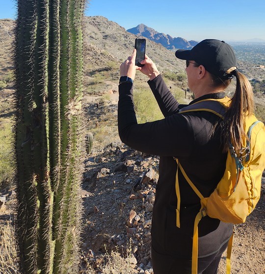 A Phoenix hiking tours guest gets up close and personal for a vacation picture with a cactus in the foreground of a beautiful Sonoran Desert landscape.