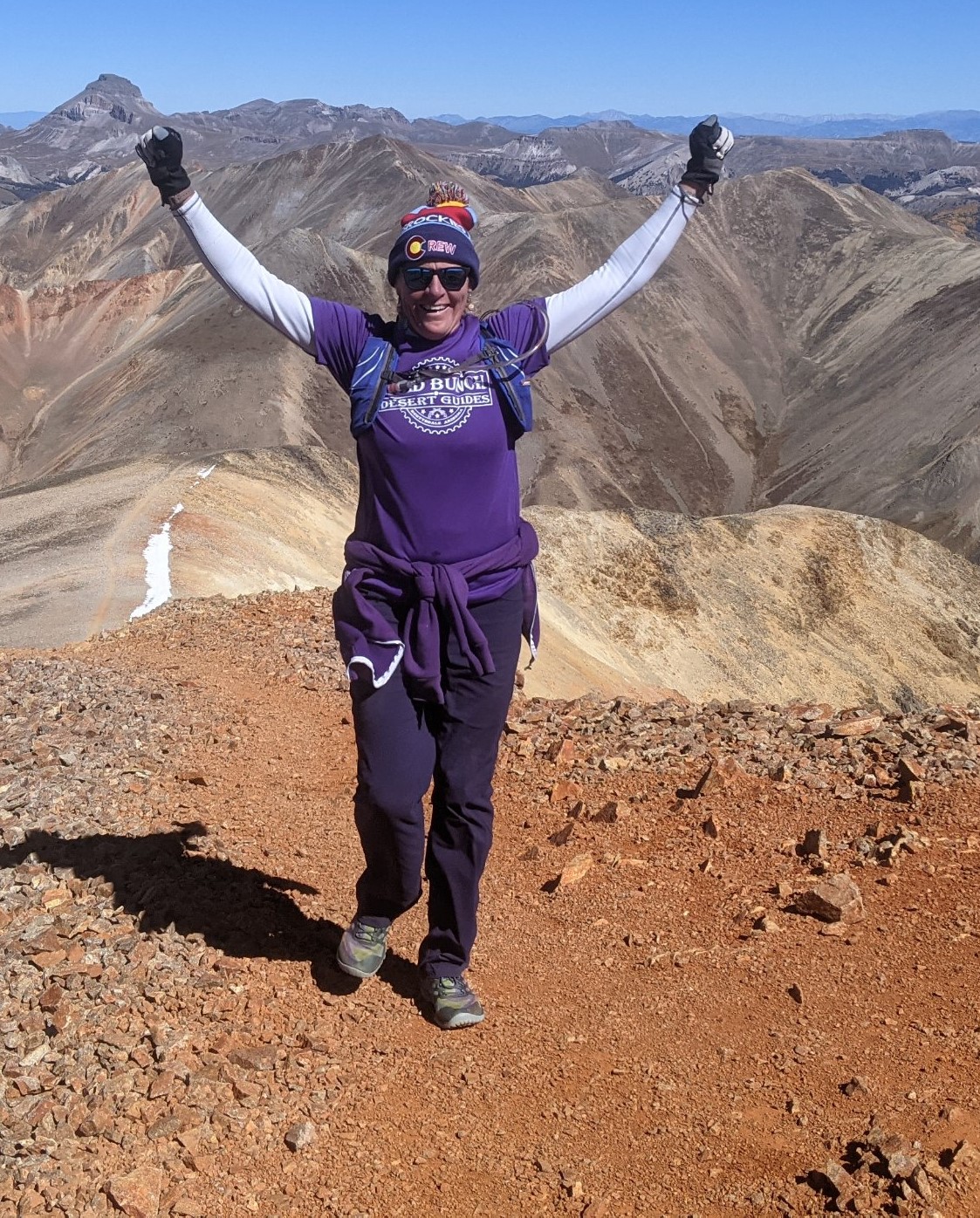 Arms raised, Laurel Darren celebrates reaching the summit of Redcloud with the vast beauty of Colorado's mountains looming majestically behind her.