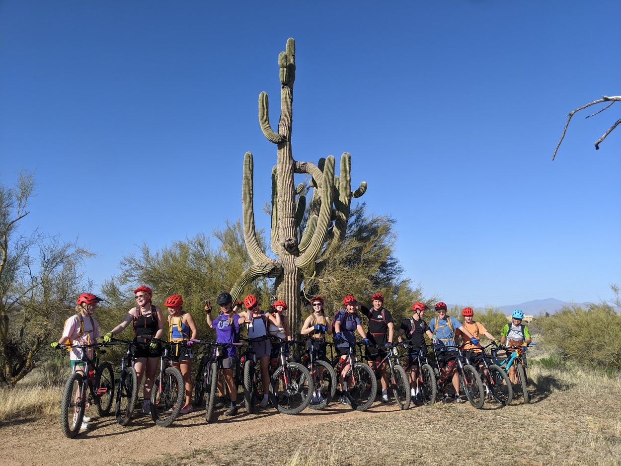 A large group from the Wild Bunch Desert Guides takes a break from a Phoenix mountain bike tour to take a picture together in front of an iconic Saguaro Cactus.